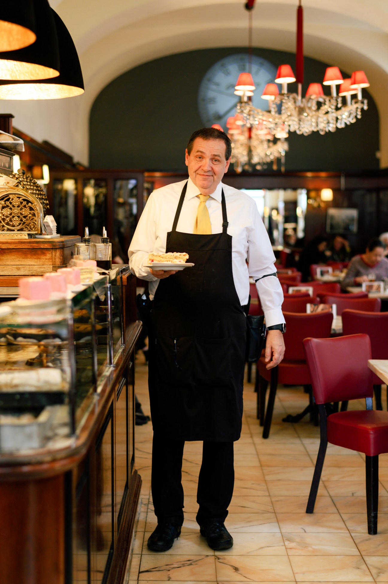 Hans Diglas in white shirt and black apron holding a plate of cake in his Vienna café
