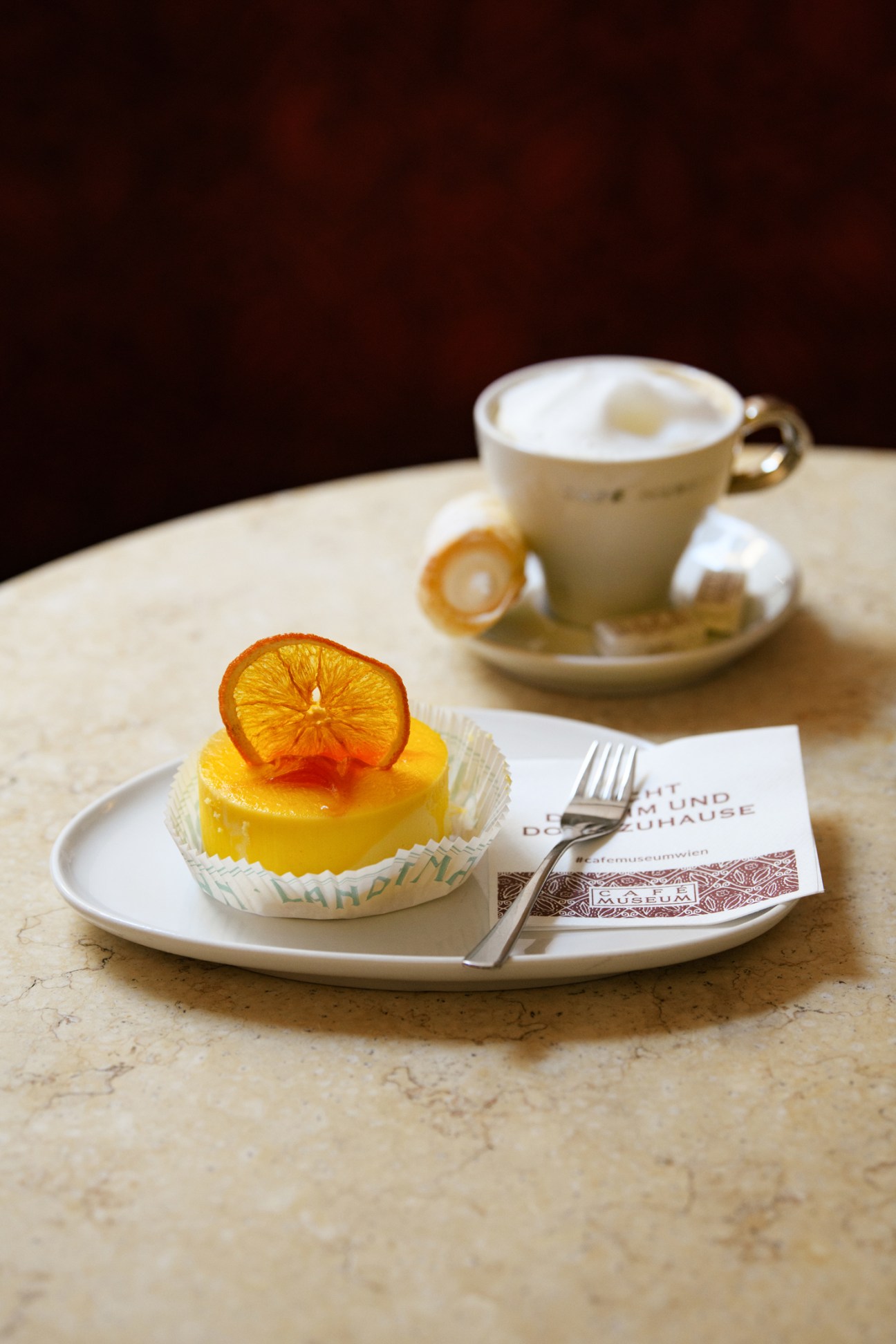 Small yellow lemon cheesecake with dried orange slice garnish on white plate with fork and coffee cup