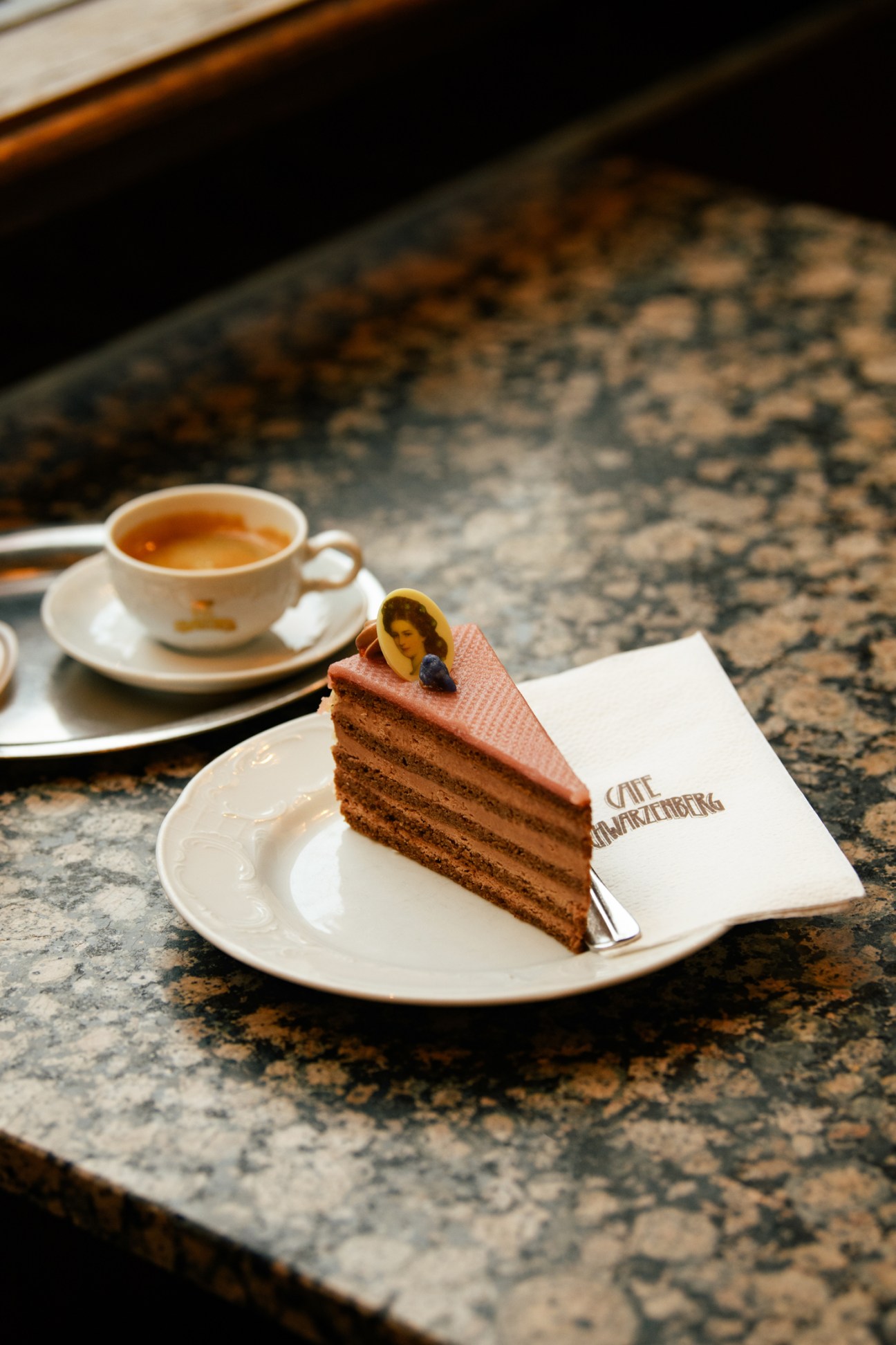 Triangular slice of layered Sissi torte with pink icing and decorative topper on white plate beside coffee cup