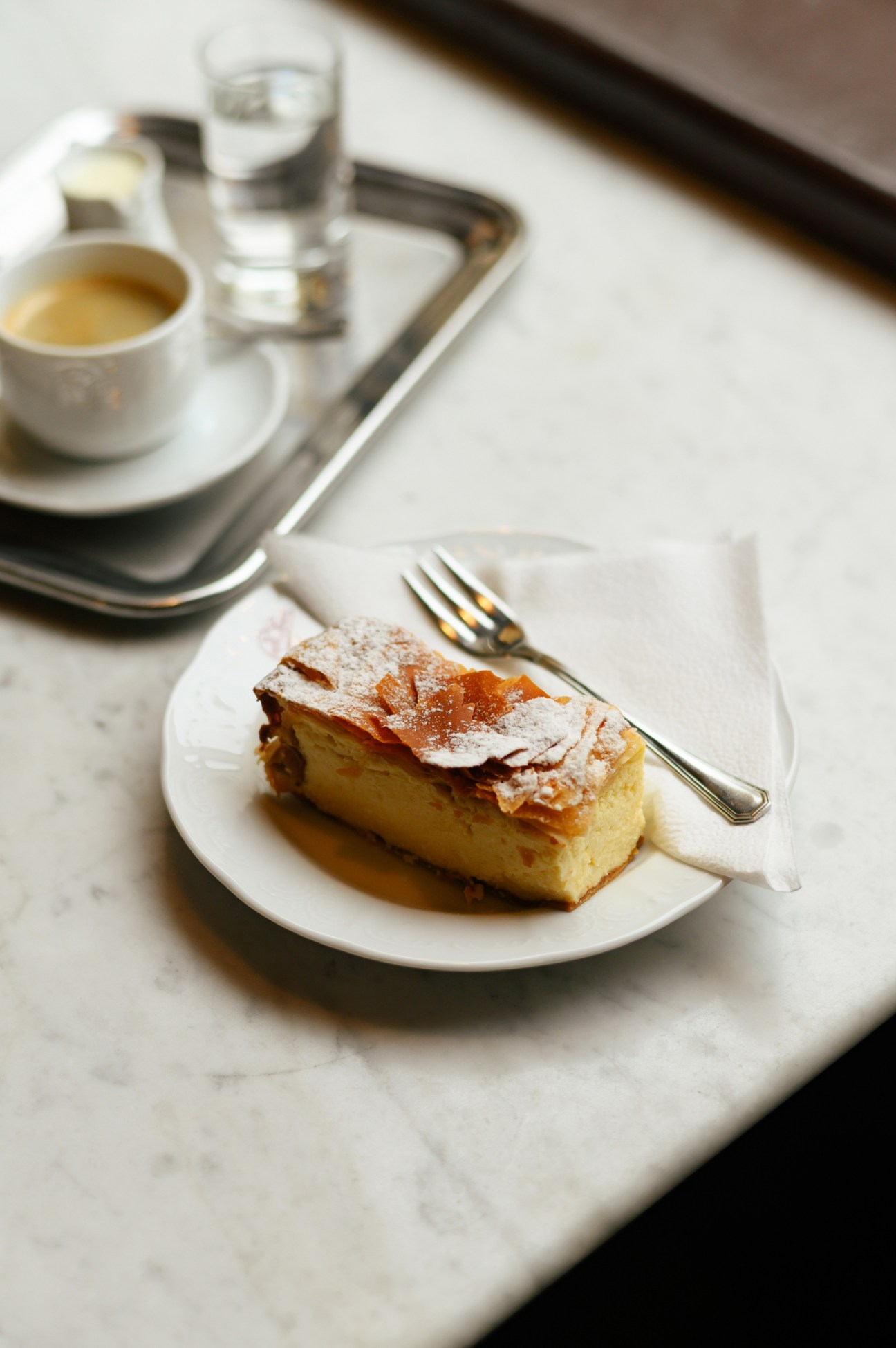 Slice of Topfenstrudel on white plate with fork and coffee cup on tray in background