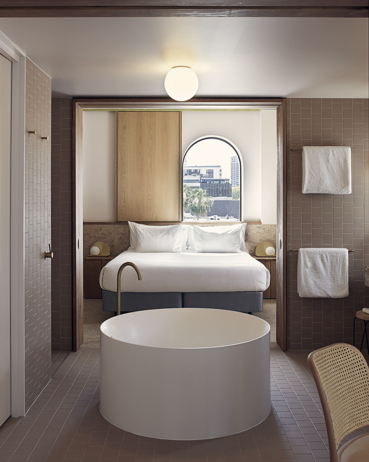 Modern hotel bathroom with round white soaking tub, bed visible through open doorway, and city view through arched window.