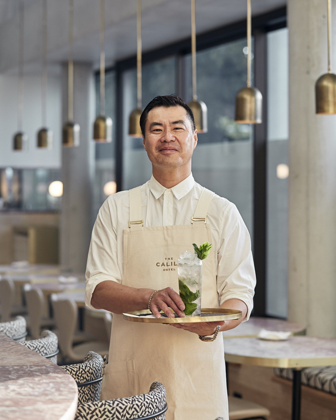 Staff member at The Calile hotel holding a tray in an elegant dining space with brass pendant lights.