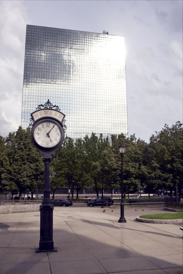 A vintage street clock in Newark's Military Park with a modern glass skyscraper and trees in the background.