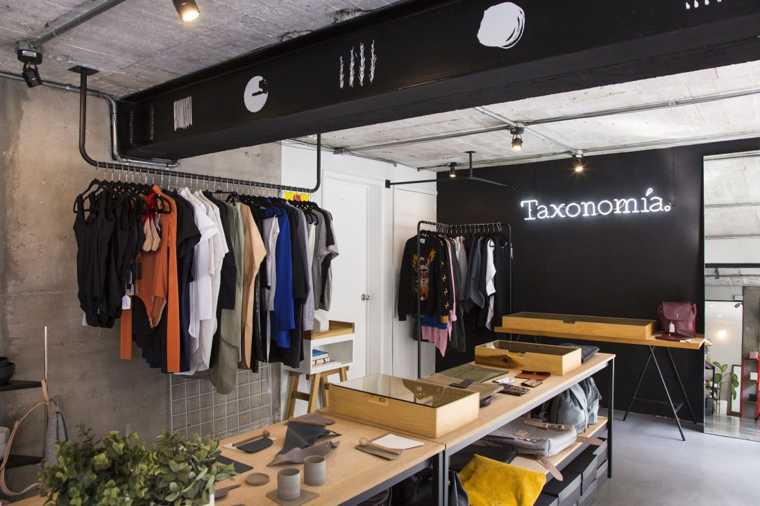 Taxonomía shop interior with clothing racks, wooden display tables, and neon signage on black wall.