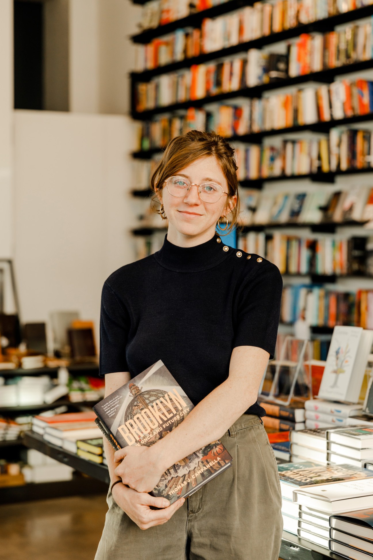 Woman with glasses holding books stands in front of blurred bookshelves at The Center for Fiction.