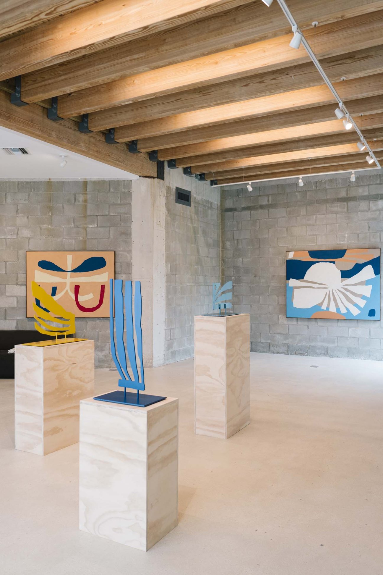 Contemporary art gallery space with wooden beam ceiling, concrete walls, colorful abstract artworks, and sculptural installations on pedestals.