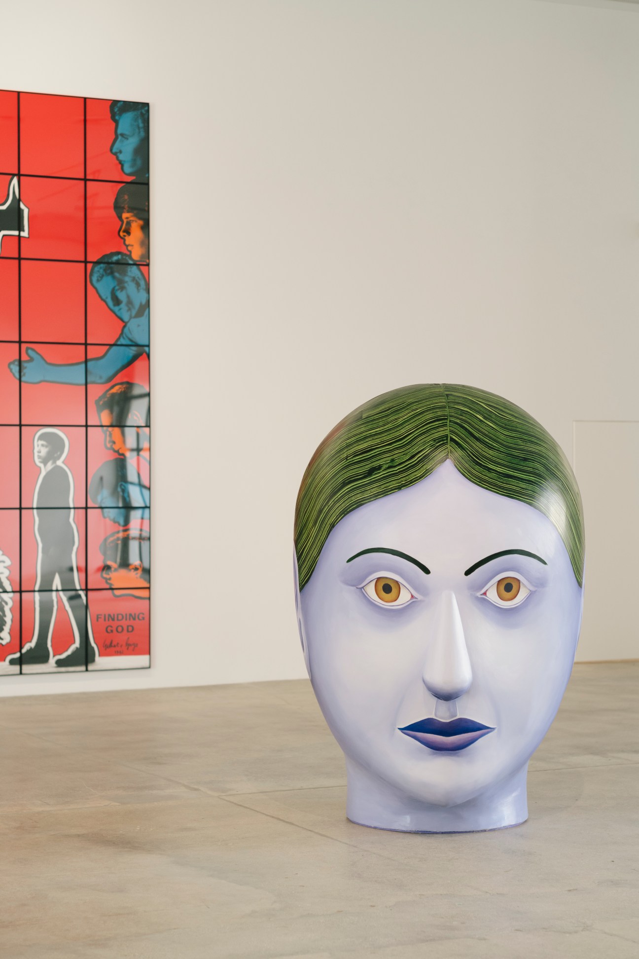 Large sculptural head with pale blue skin, green hair, yellow eyes and blue lips displayed in a white gallery space