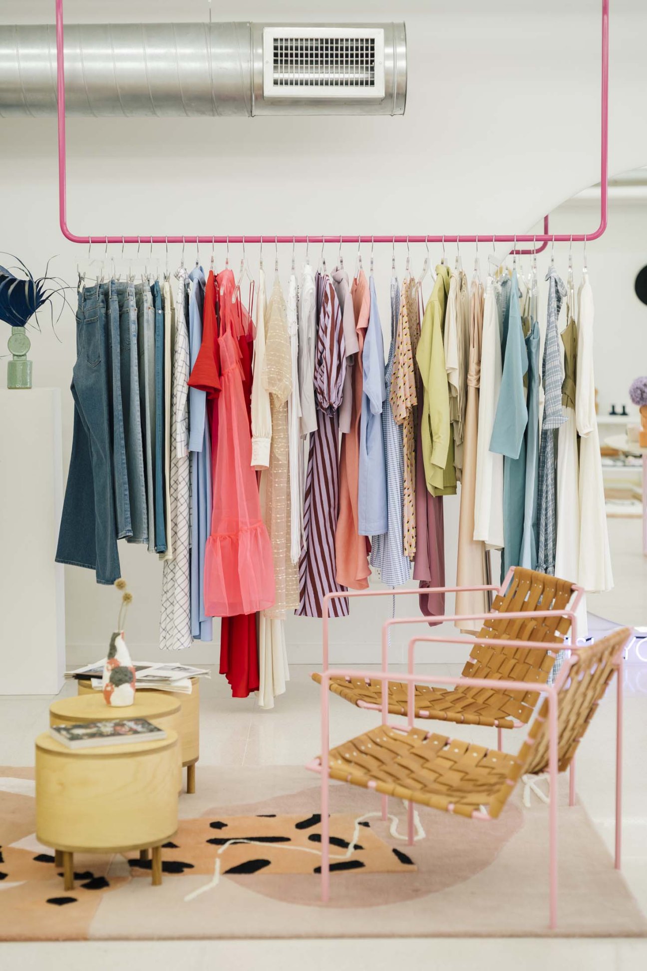 Colorful clothing displayed on a pink metal rack in a minimalist retail space with wooden chairs and yellow furnishings.