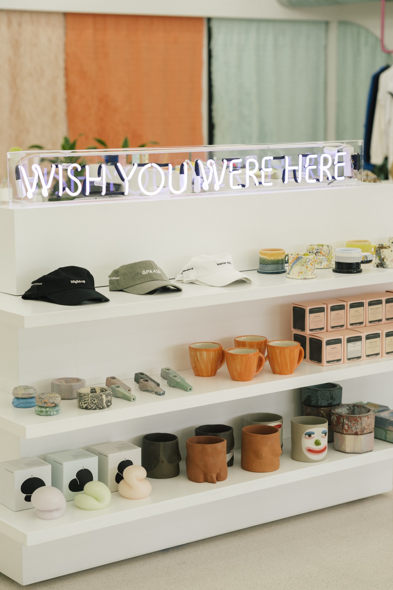 White shelving displaying ceramics, wooden bowls, hats, and accessories with neon 'Wish You Were Here' sign above.