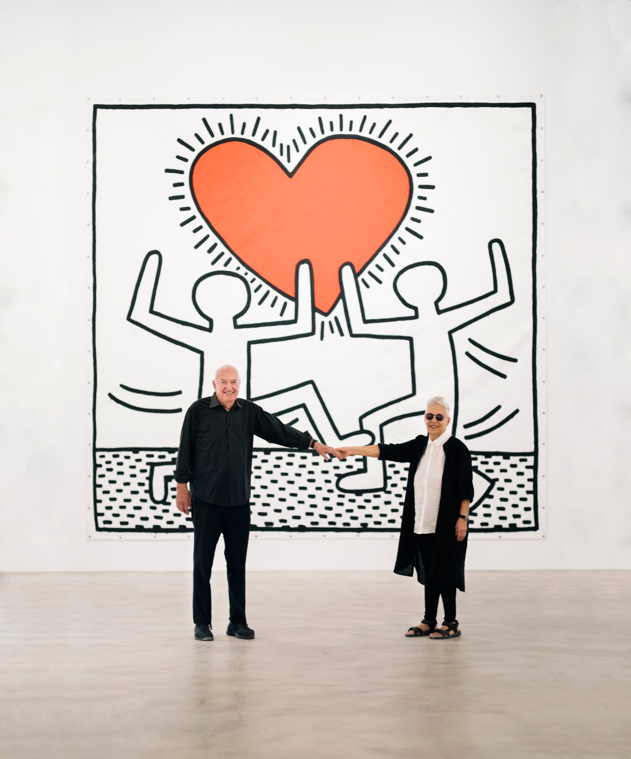 Two people standing and holding hands in front of a large Keith Haring artwork with dancing figures and a heart