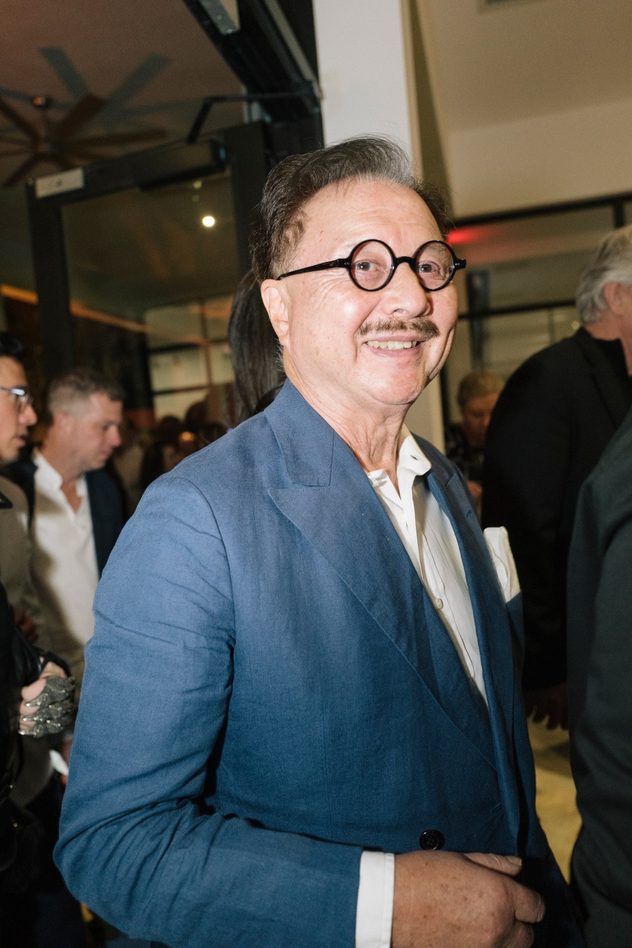 Los Angeles restaurateur Michael Chow smiling at an art gallery opening wearing a blue suit and round glasses