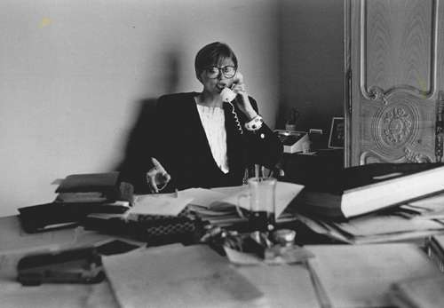 Woman in 1980s power suit with shoulder pads sitting at desk on telephone.