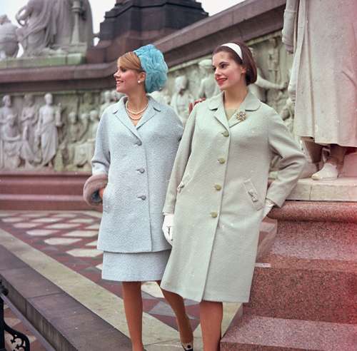 Two women in 1960s office attire wearing A-line coats, pillbox hats and kitten heels on classical steps.
