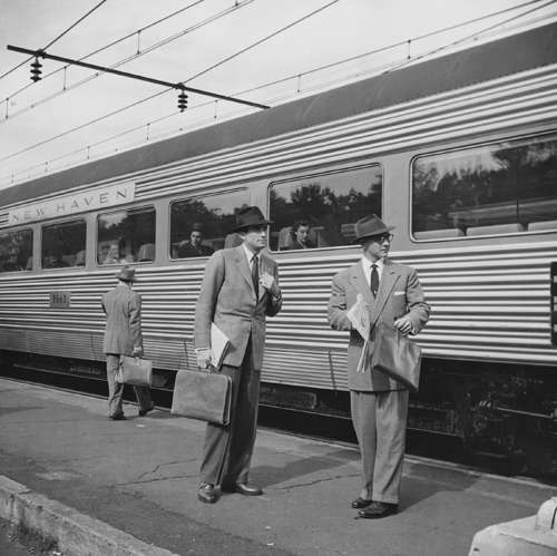 Black and white photograph of two men in gray suits with hats standing beside a train platform.