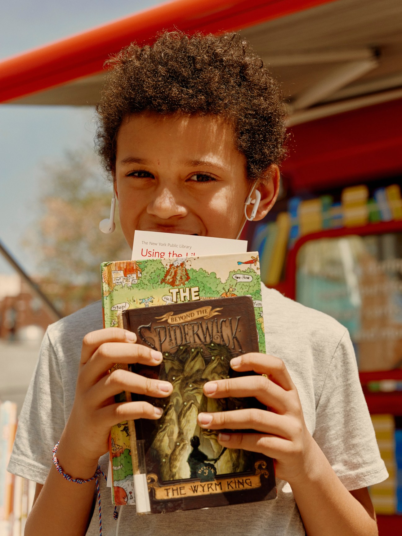 Eight-year-old Aidan holds books including 'The Spiderwick Chronicles: The Wyrm King' while standing in front of the red NYPL Bookmobile.