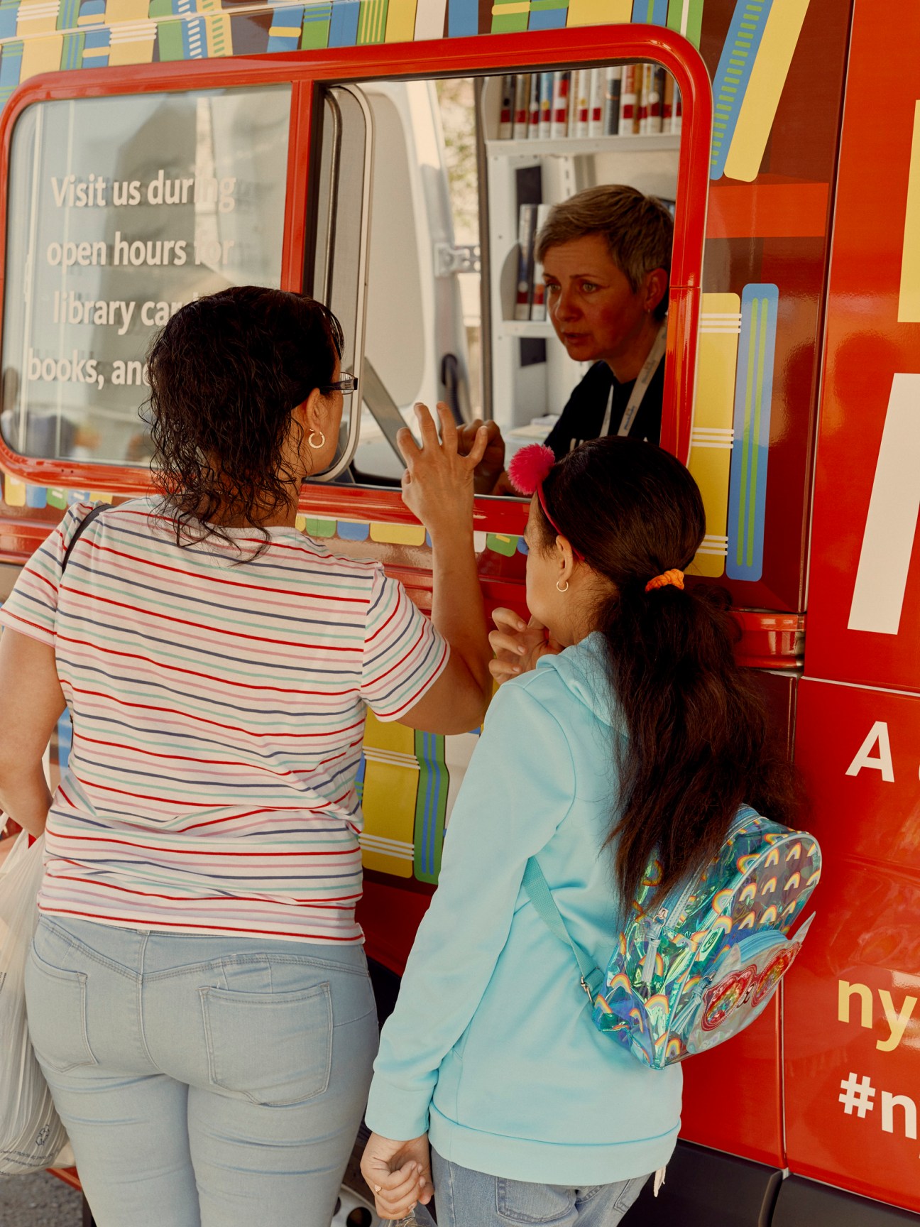 A librarian assists two young girls at the service window of the red NYPL Bookmobile.