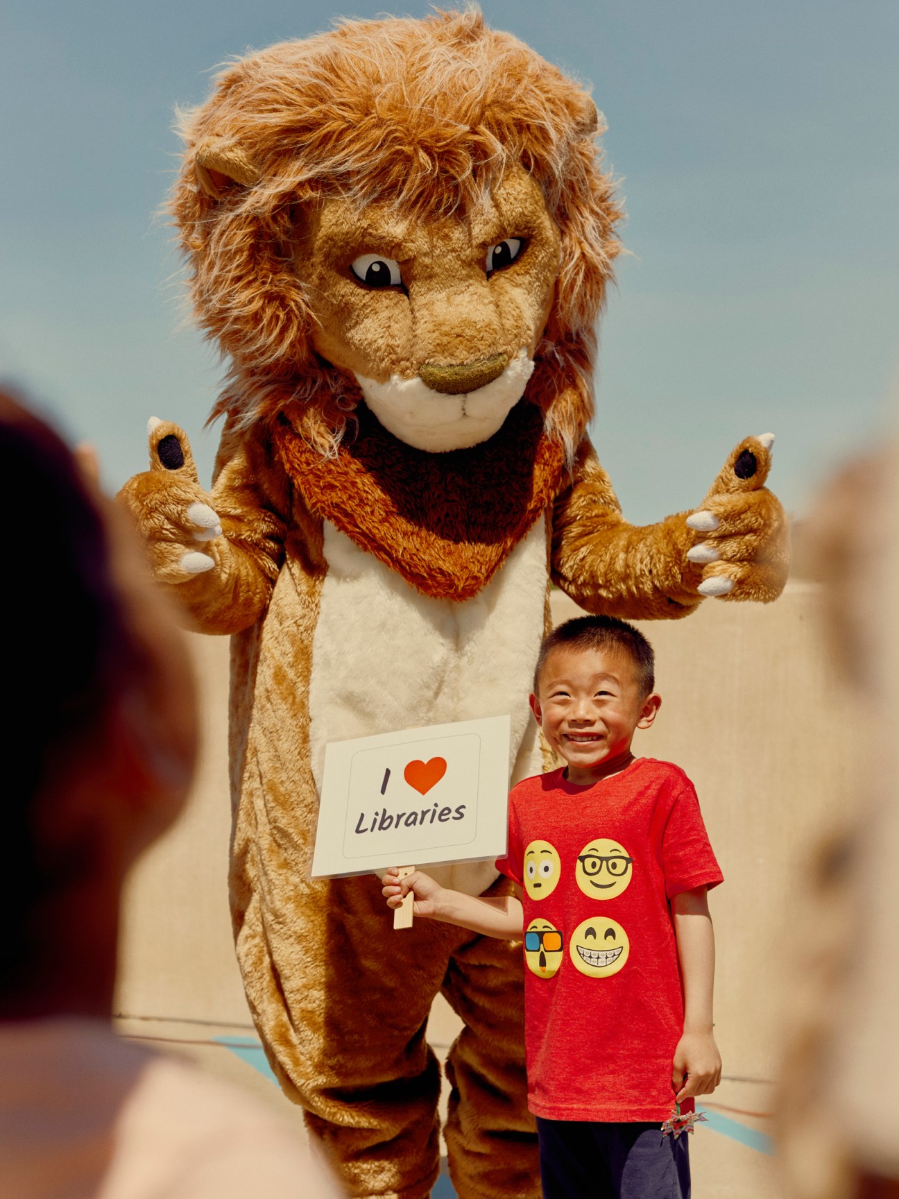 A lion mascot holds an "I love Libraries" sign while posing with a smiling young child in a red shirt.