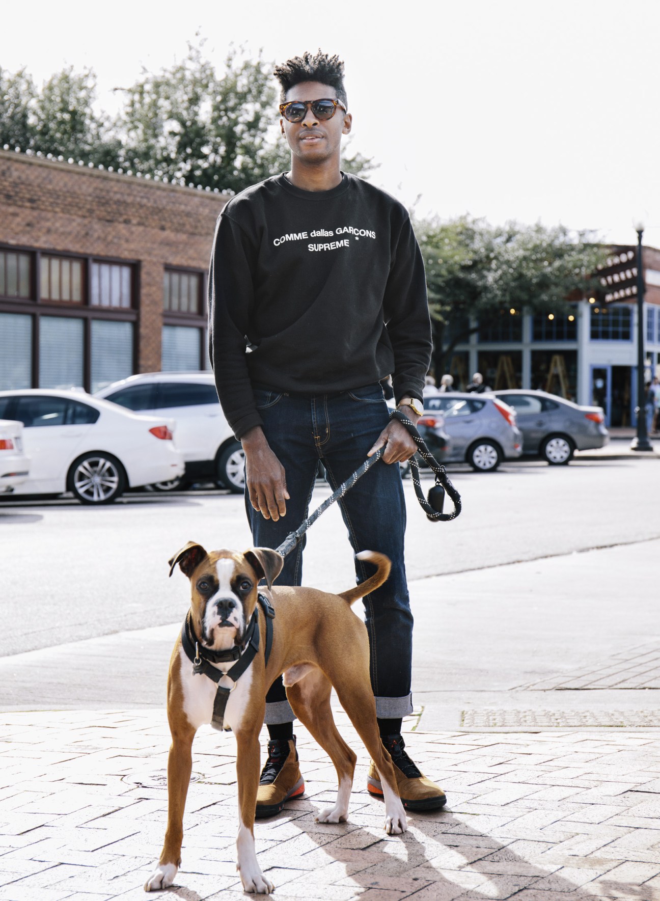 Man in Comme des Garçons Supreme sweatshirt and sunglasses stands with a brown and white boxer dog on a city street.