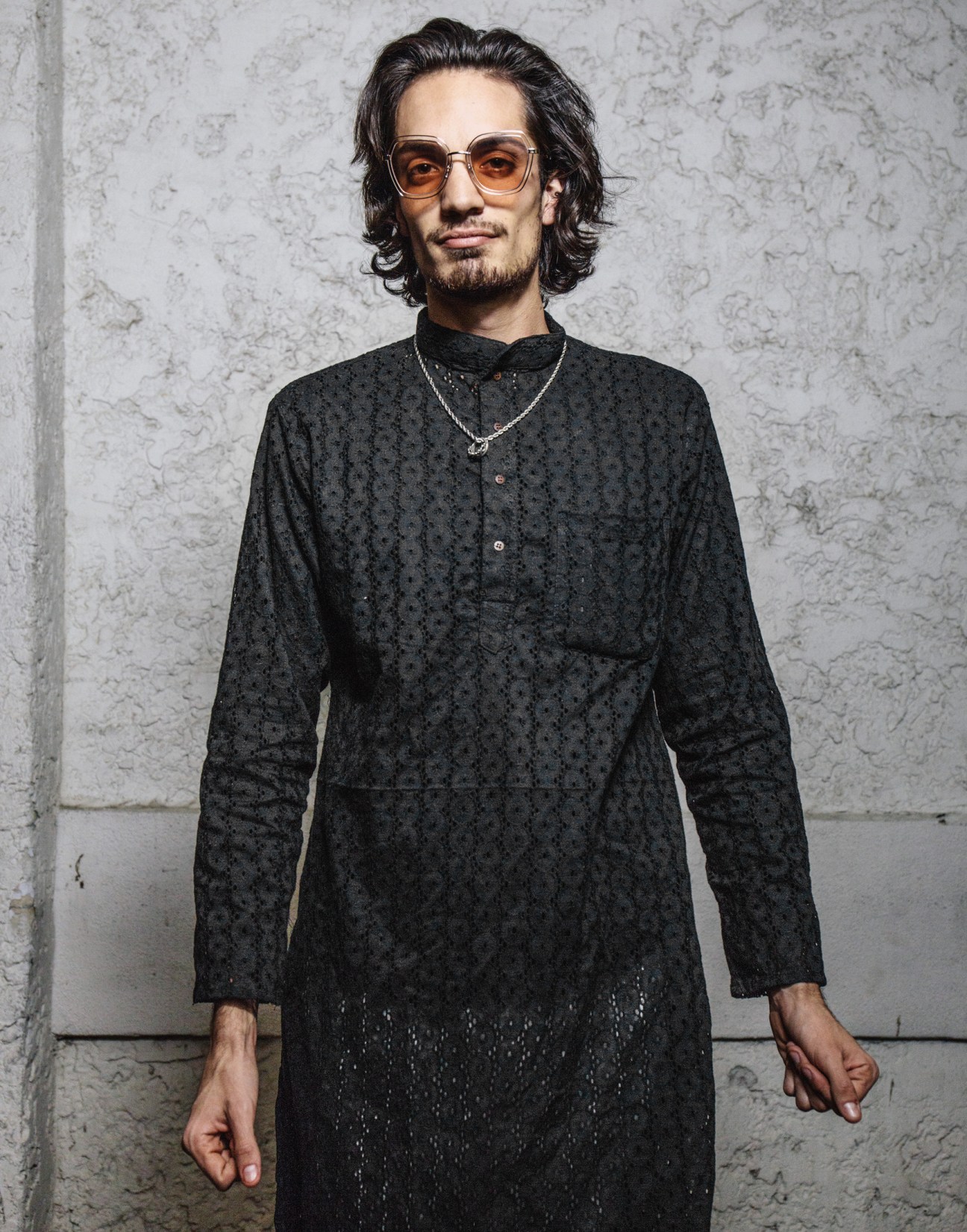 Man wearing black patterned shirt and sunglasses against textured wall background.