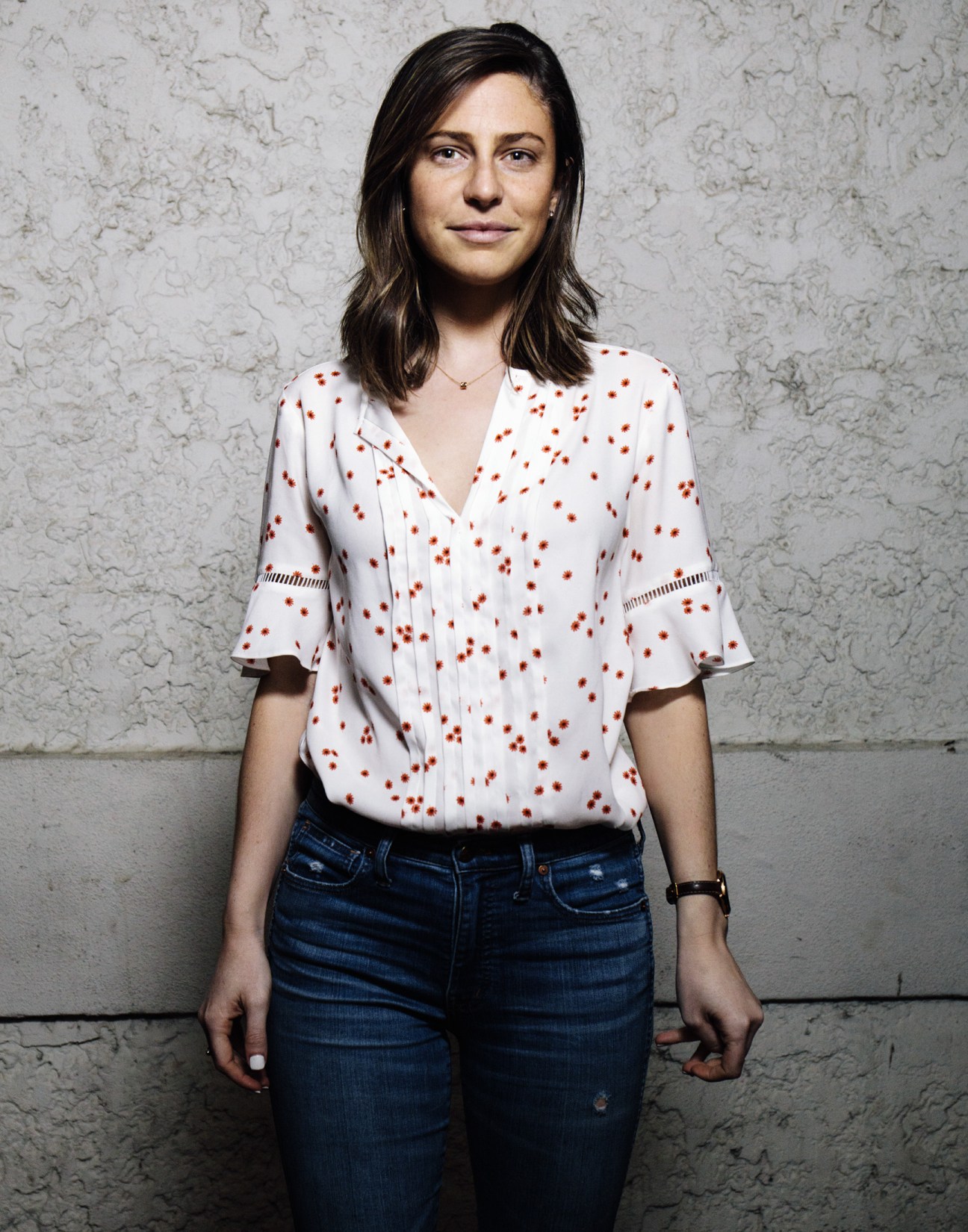Woman in white patterned blouse and jeans against textured wall.