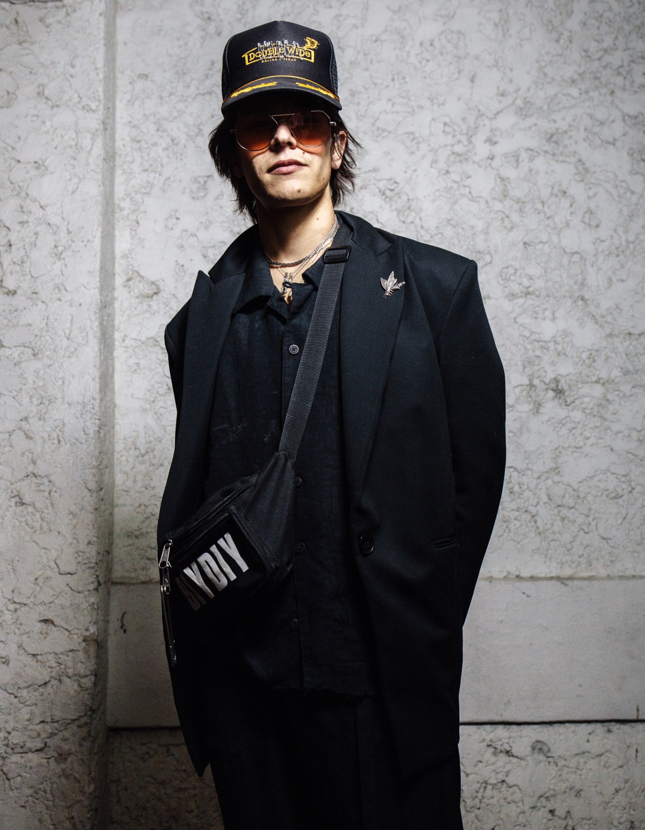 Person in black blazer, cap, and sunglasses poses against concrete wall in Dallas.