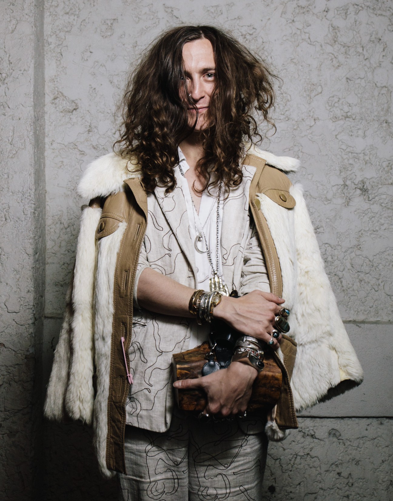 Person with long dark curly hair wearing a cream patterned shirt, tan and white fur-trimmed jacket, and multiple rings and necklaces at a Dallas venue.
