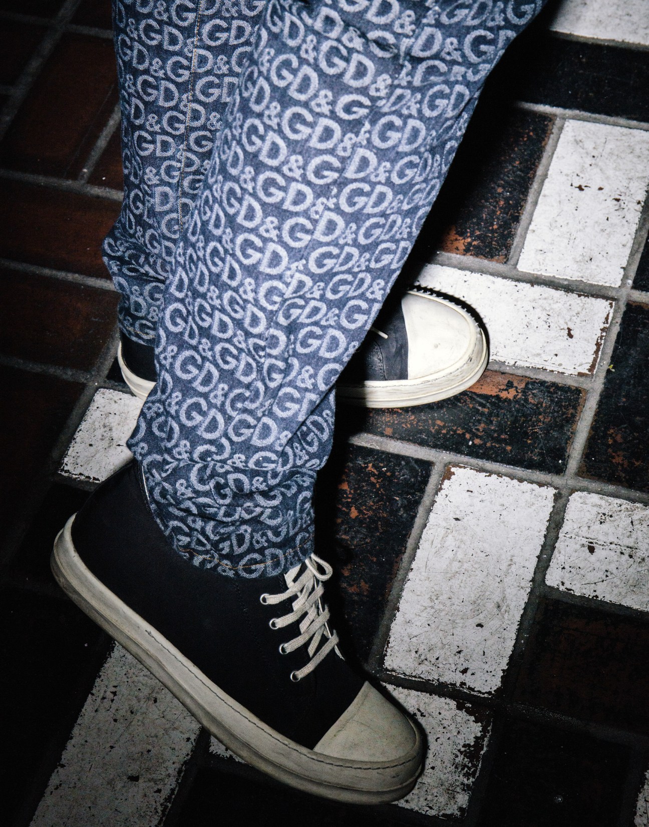 Person wearing patterned blue jeans and white canvas sneakers standing on a tiled floor.
