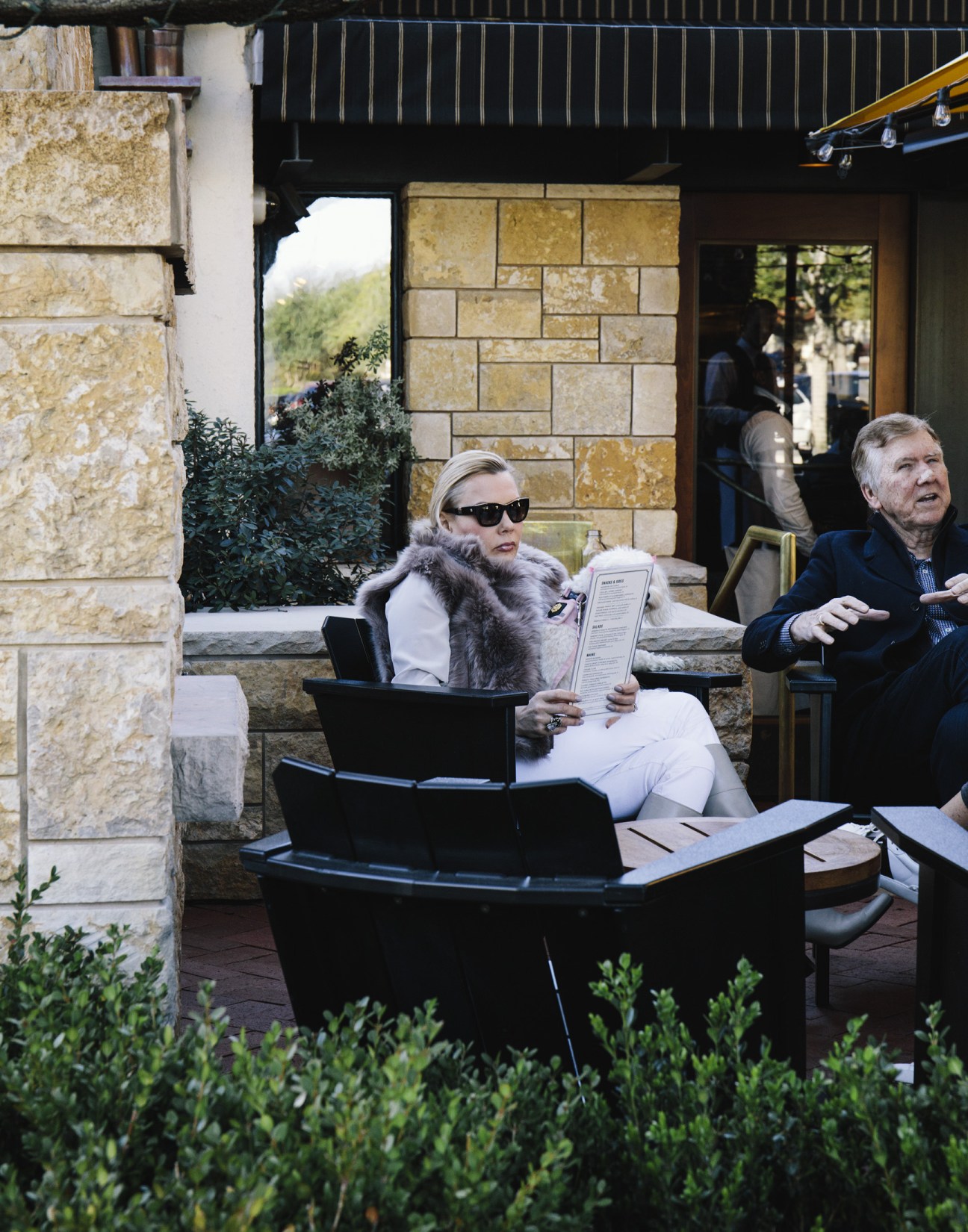 Woman in sunglasses and fur stole holds menu at outdoor restaurant patio.