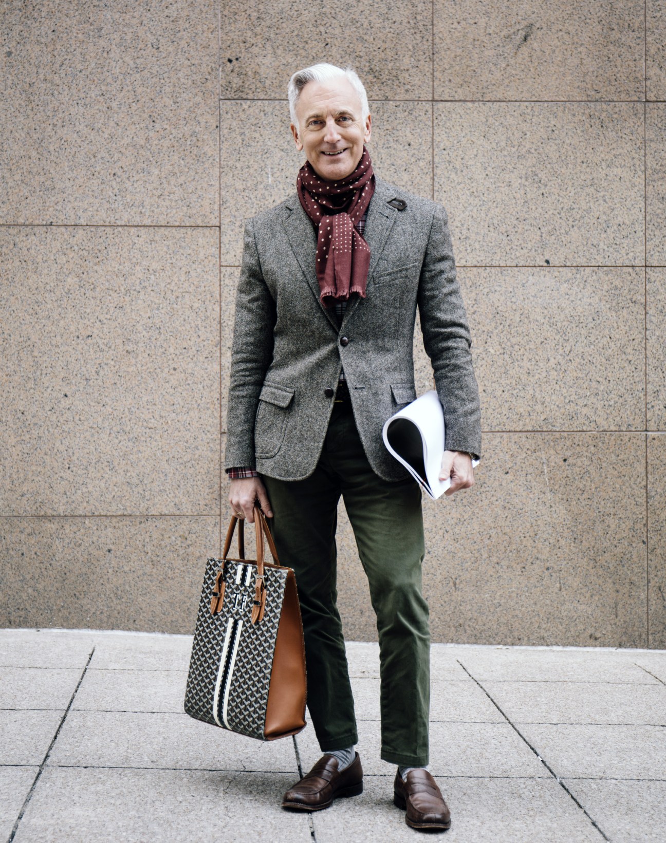 Older man in gray blazer and burgundy scarf holding a patterned tote bag stands against textured stone wall.
