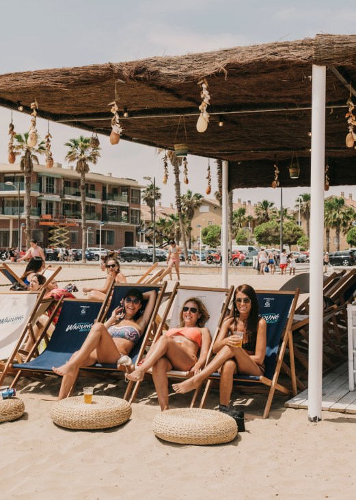 Women relaxing in deck chairs under a wooden pergola at Warung Beach chiringuito in Valencia.