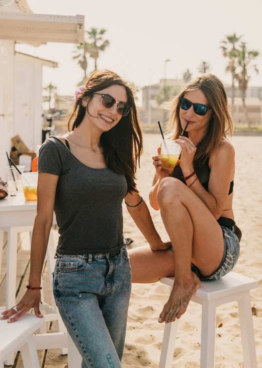 Two young women with sunglasses hold drinks with straws at a beachside chiringuito, palm trees and beach structures visible behind them.