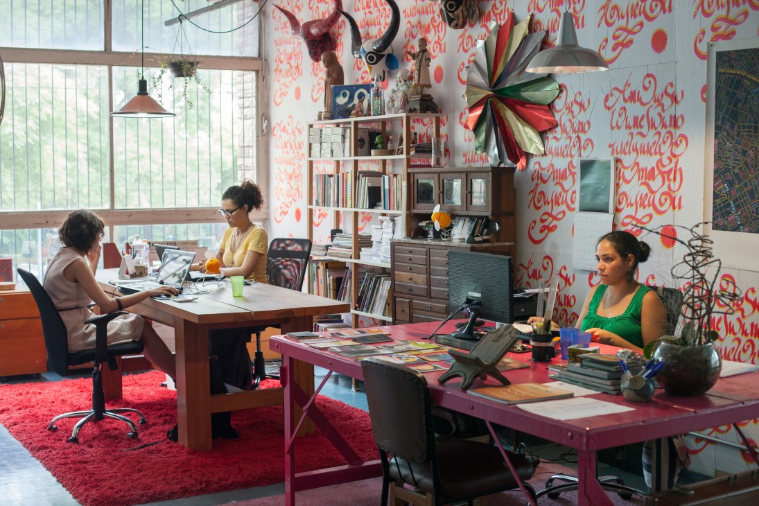 Three women work at desks in a bright office with red patterned wallpaper, shelving, and a colorful ribbon art installation.