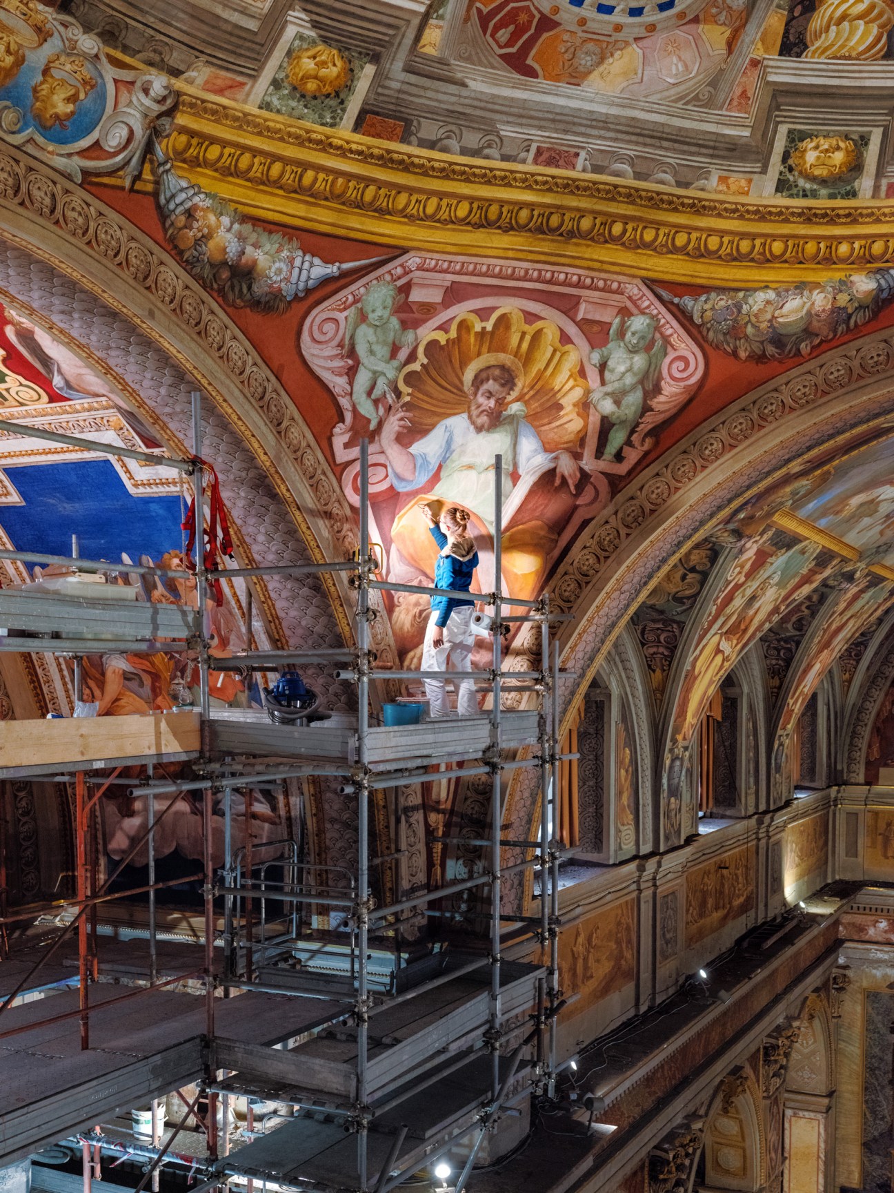 Restorer on scaffolding working on ornate baroque frescoed ceiling with religious figures and gold gilded details.