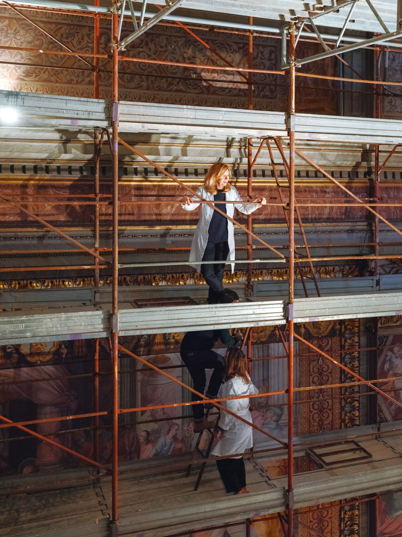 Two restorers on scaffolding examining ornate baroque frescoes and decorative wall panels in a church interior.