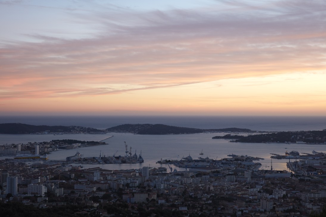 Aerial view of Toulon Bay at sunset with French naval vessels in the harbor and surrounding hills.