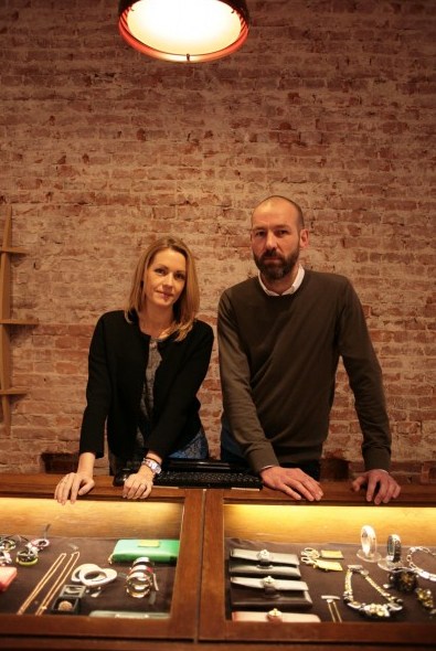 Two people stand behind a jewelry display counter in a brick-walled retail space.