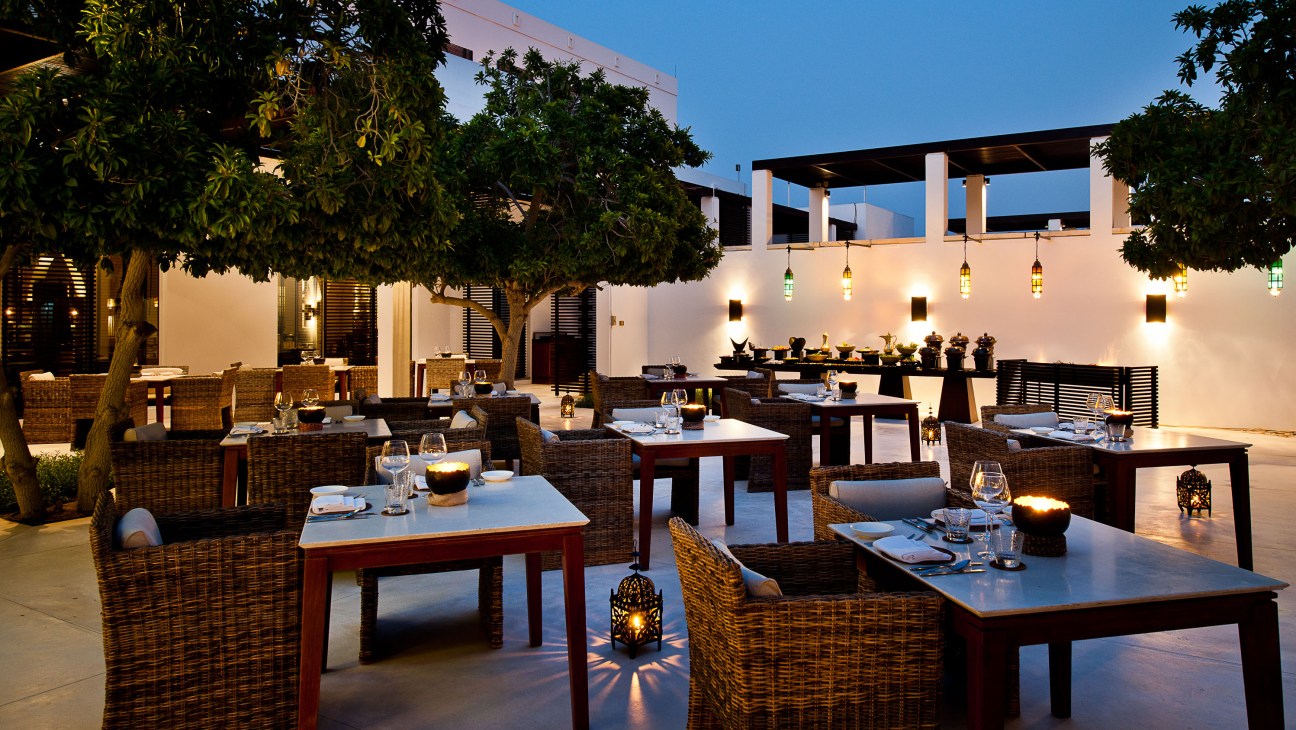 The Chedi's outdoor dining courtyard in Muscat at dusk, featuring wicker furniture, lantern lighting, and lush greenery.