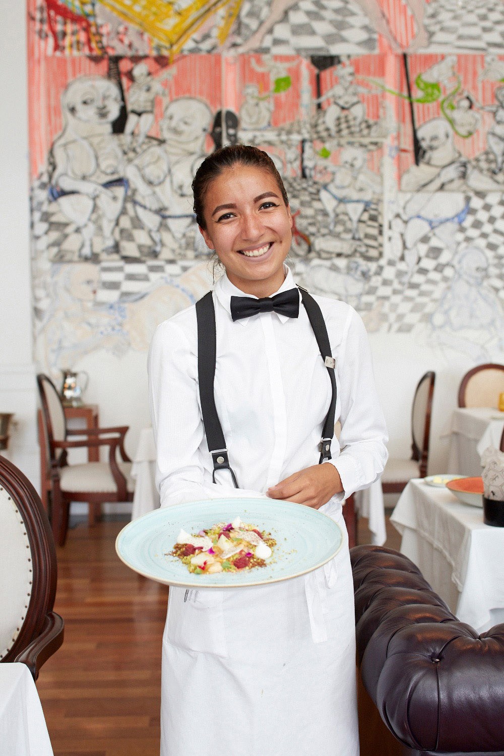 Server in formal white shirt and black bow tie holding a plated dessert in a restaurant dining room with colorful artwork on the walls.