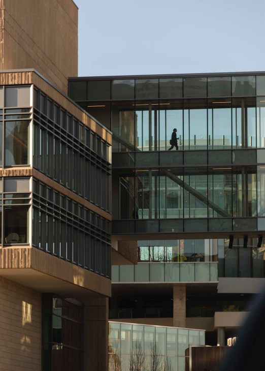 Modern office building with wood and black metal cladding and large windows, a person walking across an upper level skybridge.