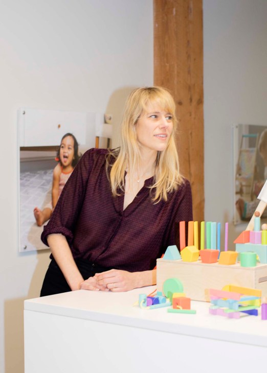 Jessica Rolph, co-founder of Lovevery, stands at a white desk with colorful educational toys displayed in front of her.