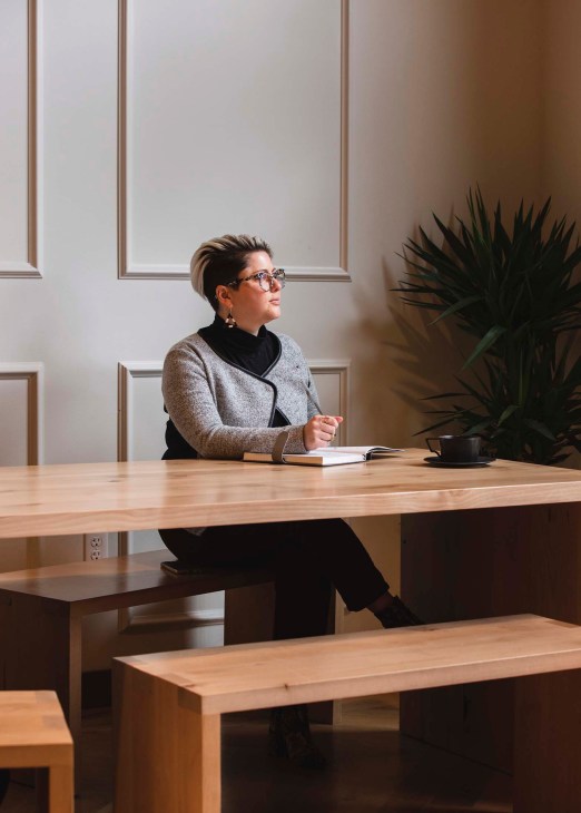 Jennifer Hensley sits at a wooden desk in a minimalist office with a notebook and coffee cup beside her.