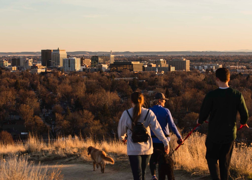 Hikers and a dog on a trail overlook Boise's skyline at sunset with forested hills in the background.