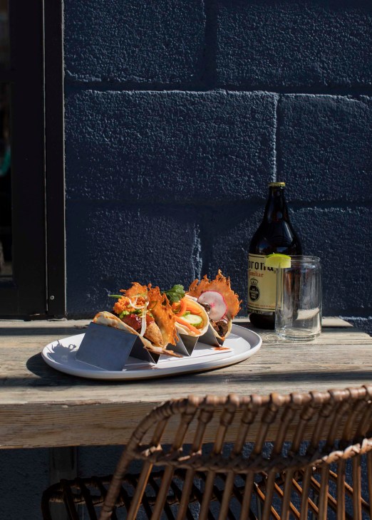 Gourmet tacos on a white plate with a beer bottle and glass against a dark brick wall.