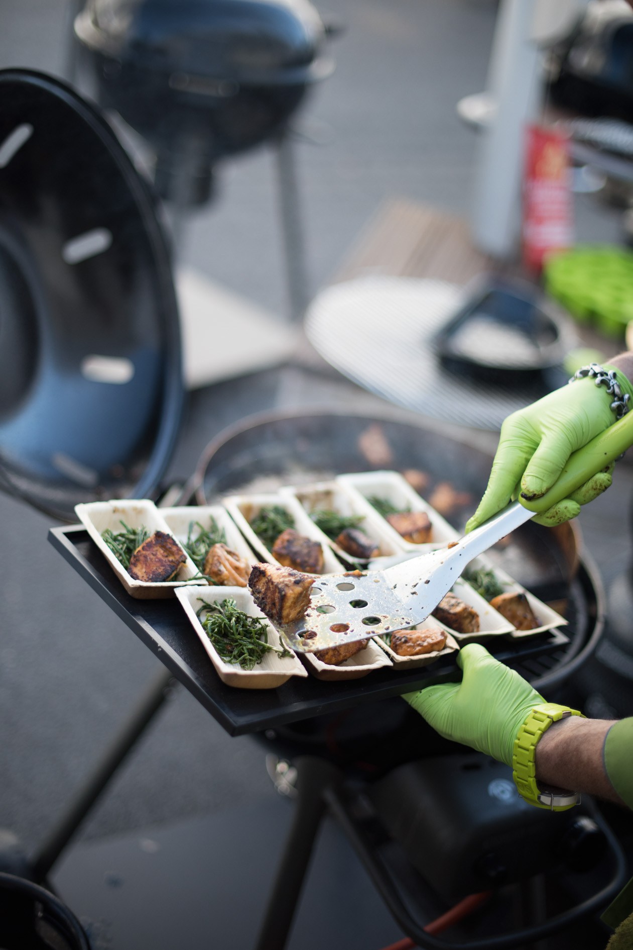 Person in bright green gloves serving grilled meat samples on white platters at the garden fair.