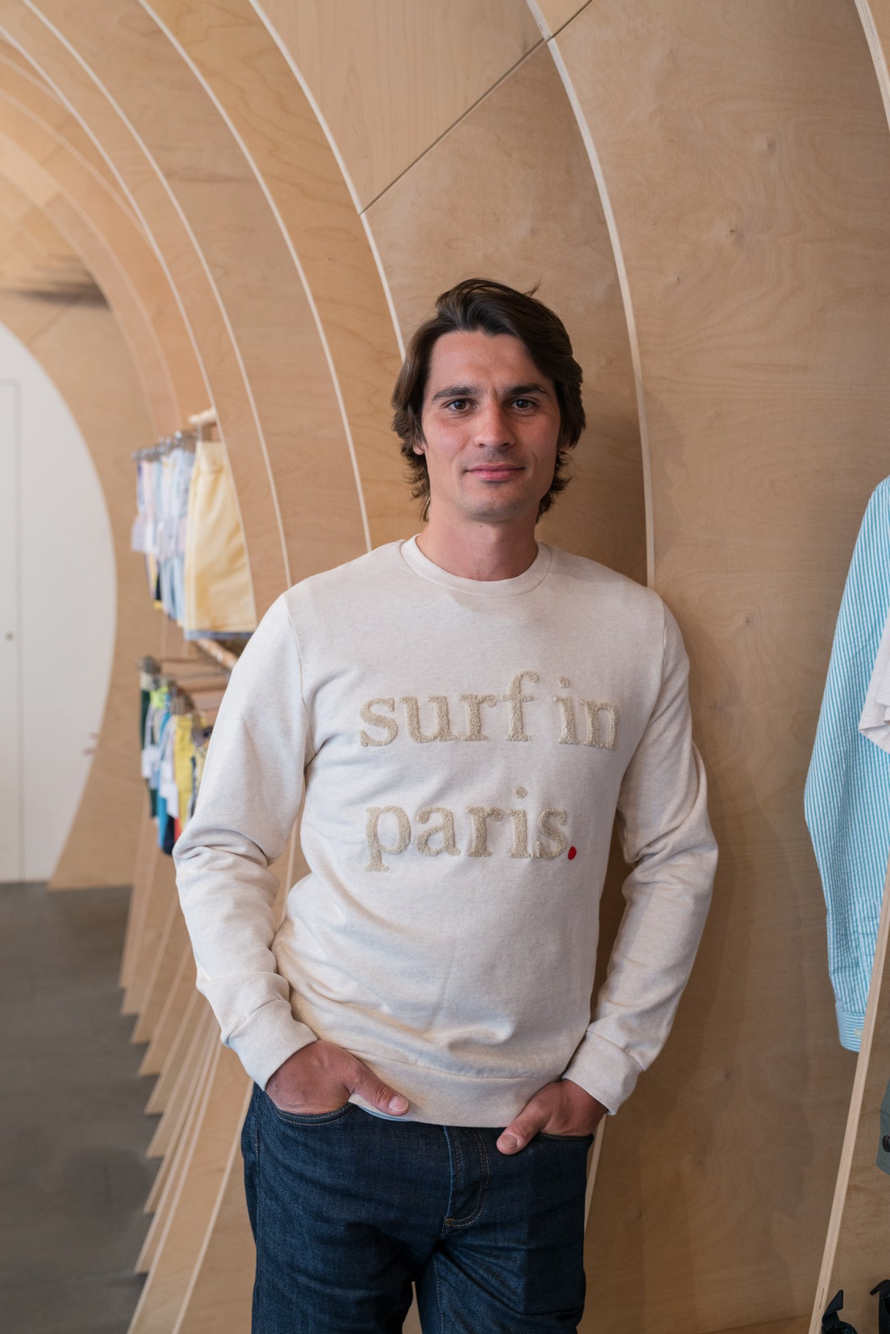 Man wearing cream sweatshirt with 'surfin paris' text in wooden interior with curved ceiling