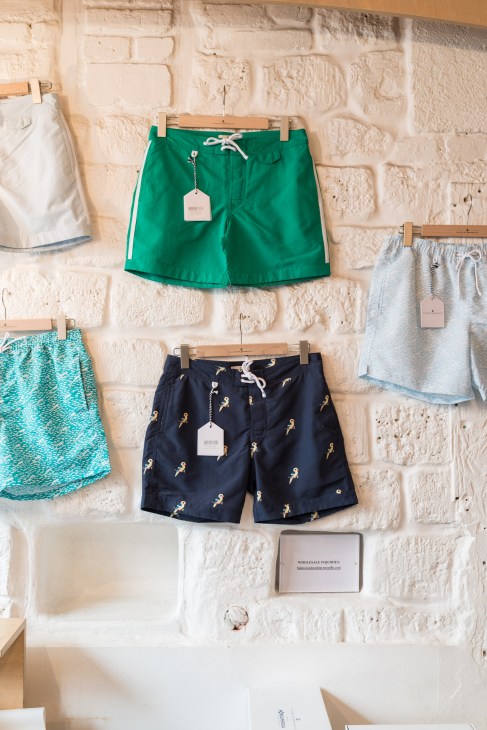 Colorful swim shorts hanging on wooden pegs against a cream brick wall, with price tags attached.