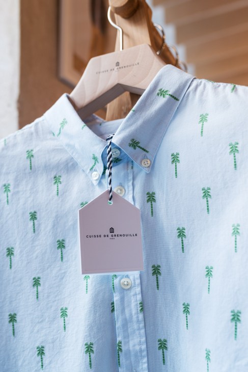 Light blue shirt with green palm tree pattern and Cuisse de Grenouille price tag on wooden hanger.