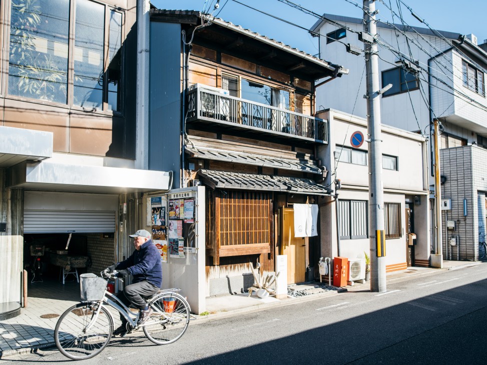 A traditional wooden machiya house exterior in Kyoto with a cyclist passing by on the street.