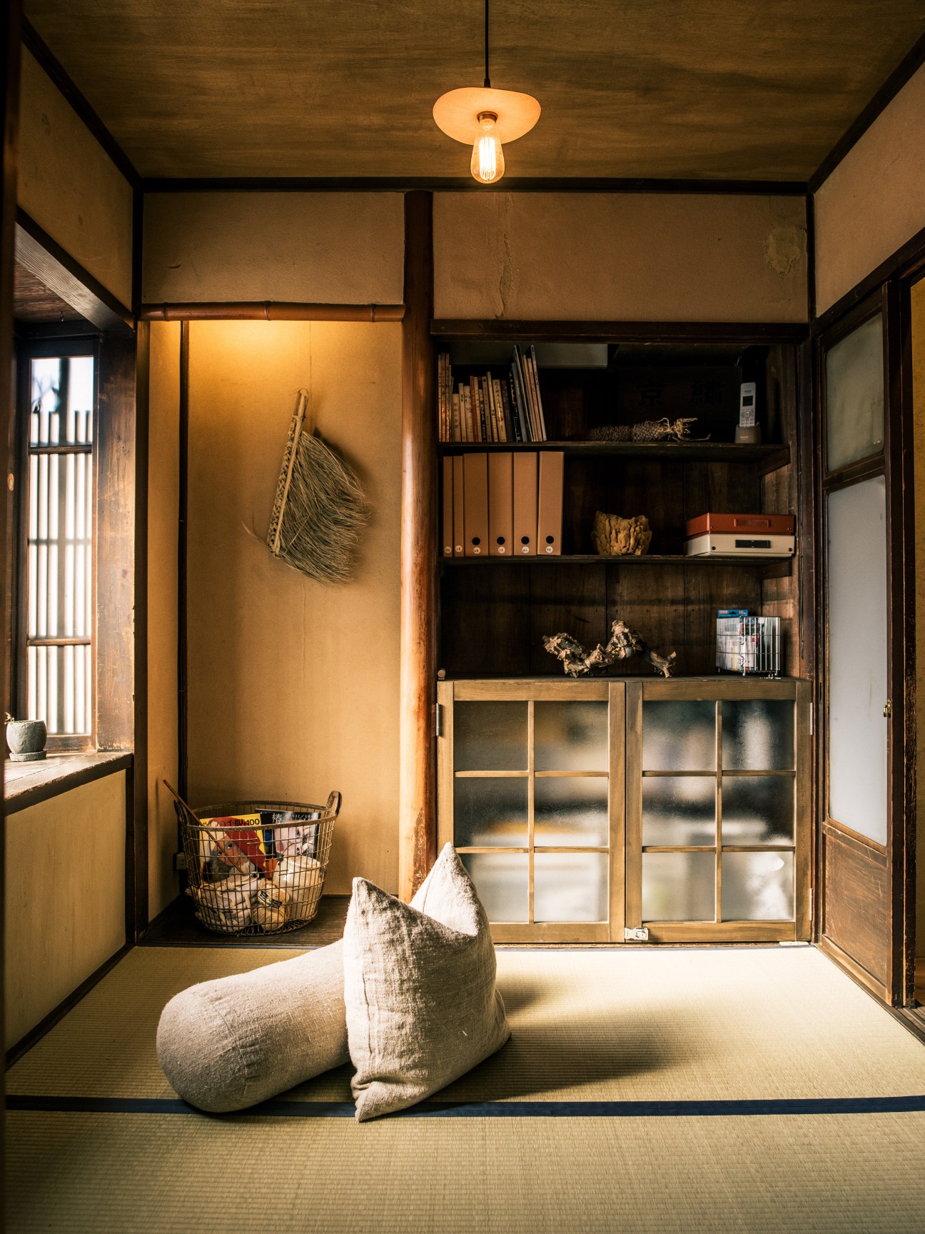 Traditional Japanese room with tatami mats, woven basket, and wooden shelving displaying books and decorative objects.