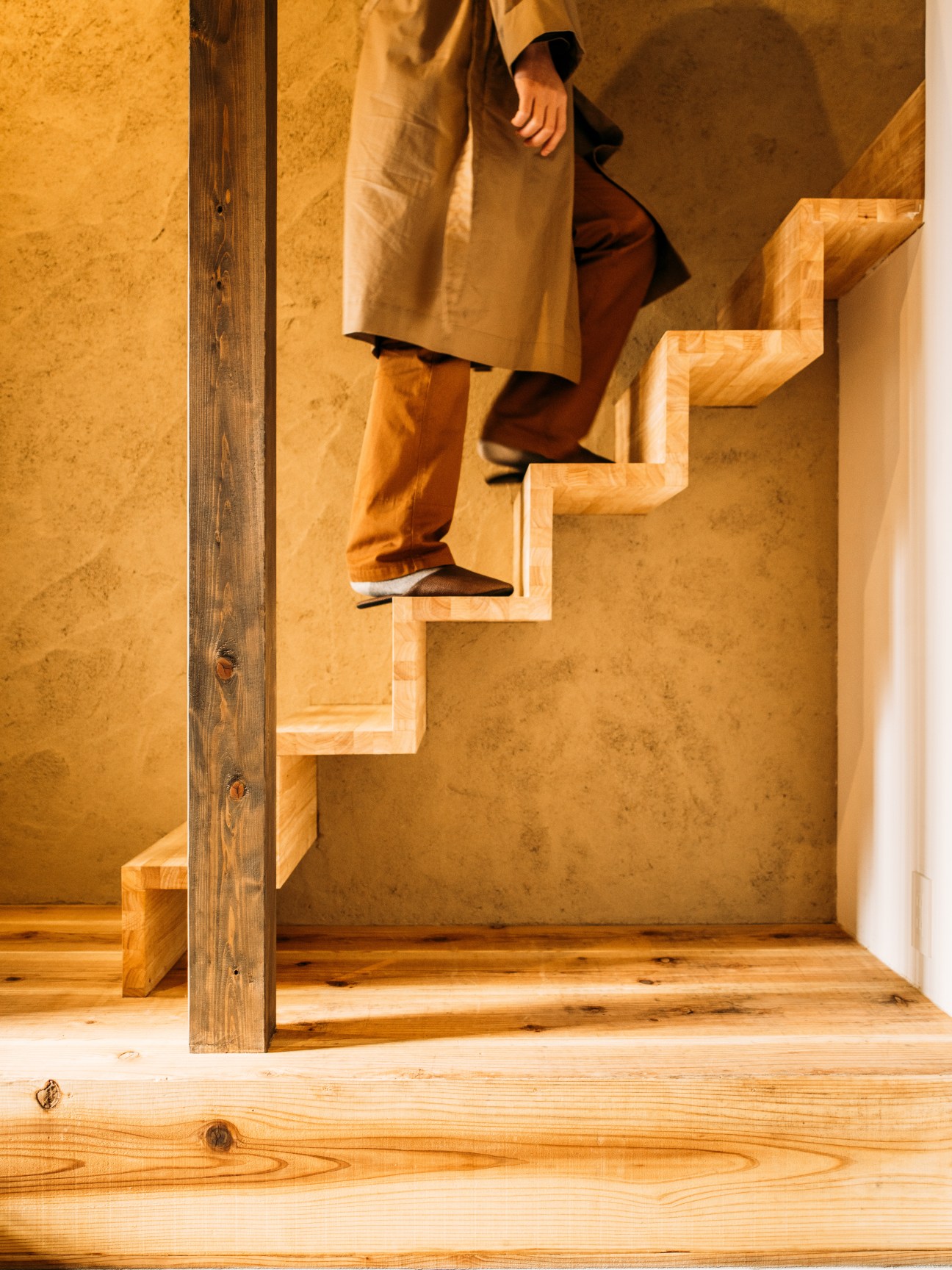 Person descending wooden floating staircase with ochre walls in renovated machiya guesthouse